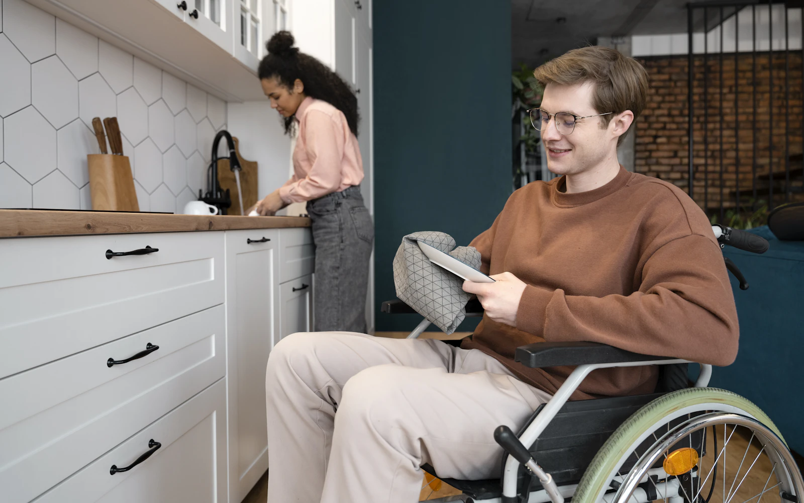 A young man in a wheelchair drying dishes in a bright kitchen while a support worker works beside him